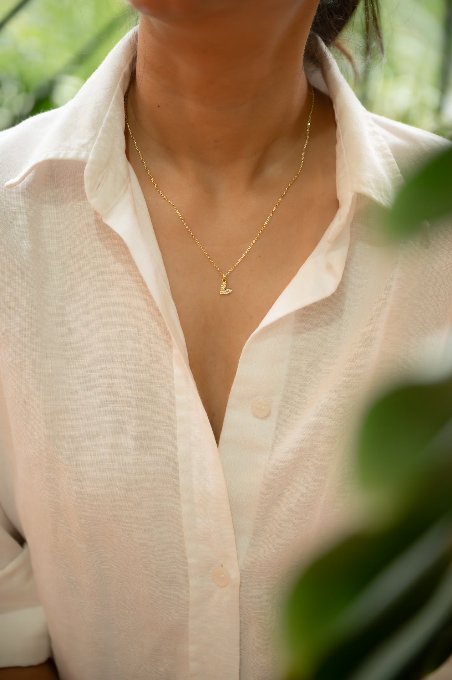 Person wearing a light-colored shirt with a gold necklace, surrounded by greenery.